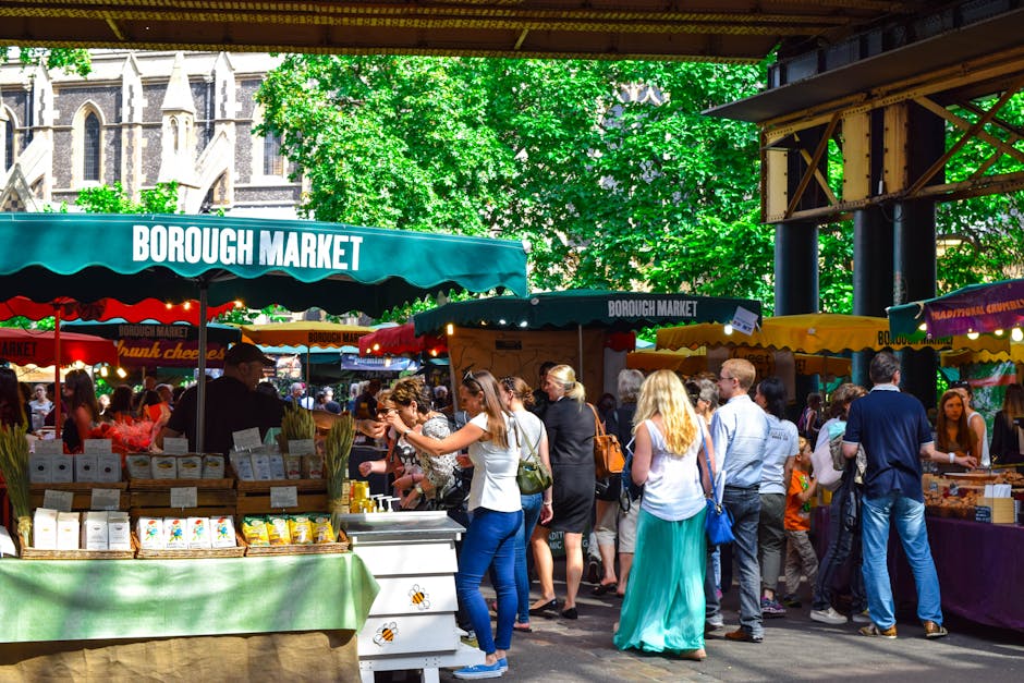 A busy outdoor scene outside Borough Market in London, with a large crowd of pedestrians walking on the pavement under a modern, multi-level metallic and glass building structure displaying the sign '